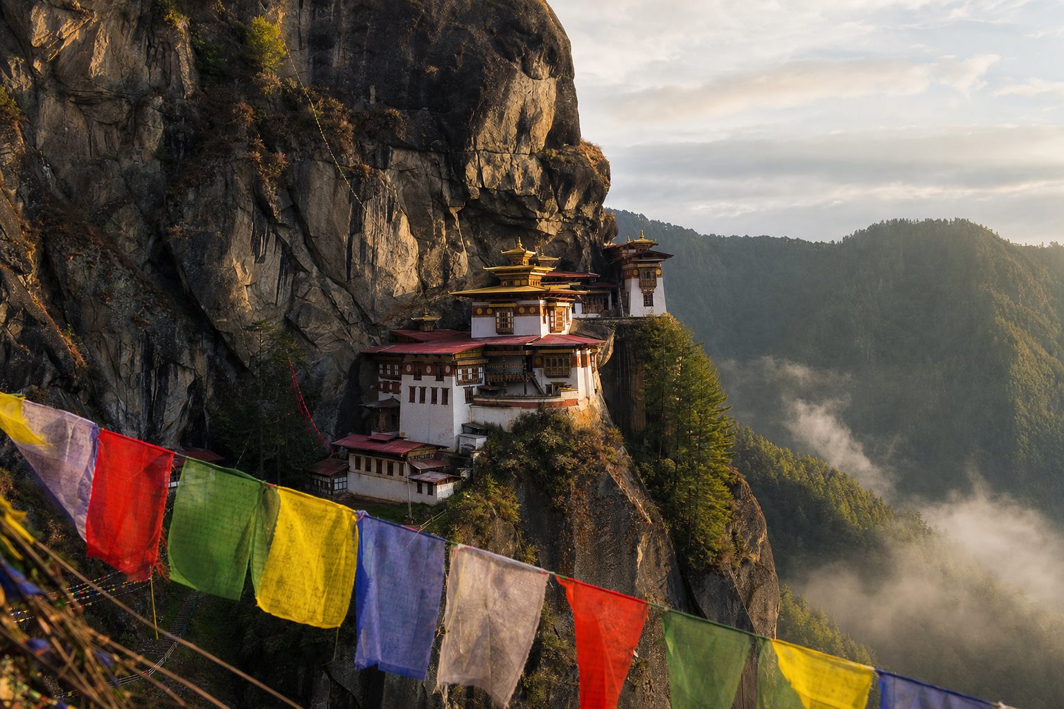 A stone path rises through pine forest in the foreground toward Tiger’s Nest monastery clinging to a sheer cliff in the midground, with misty Paro Valley ridges receding beneath soft early-morning light, no people.