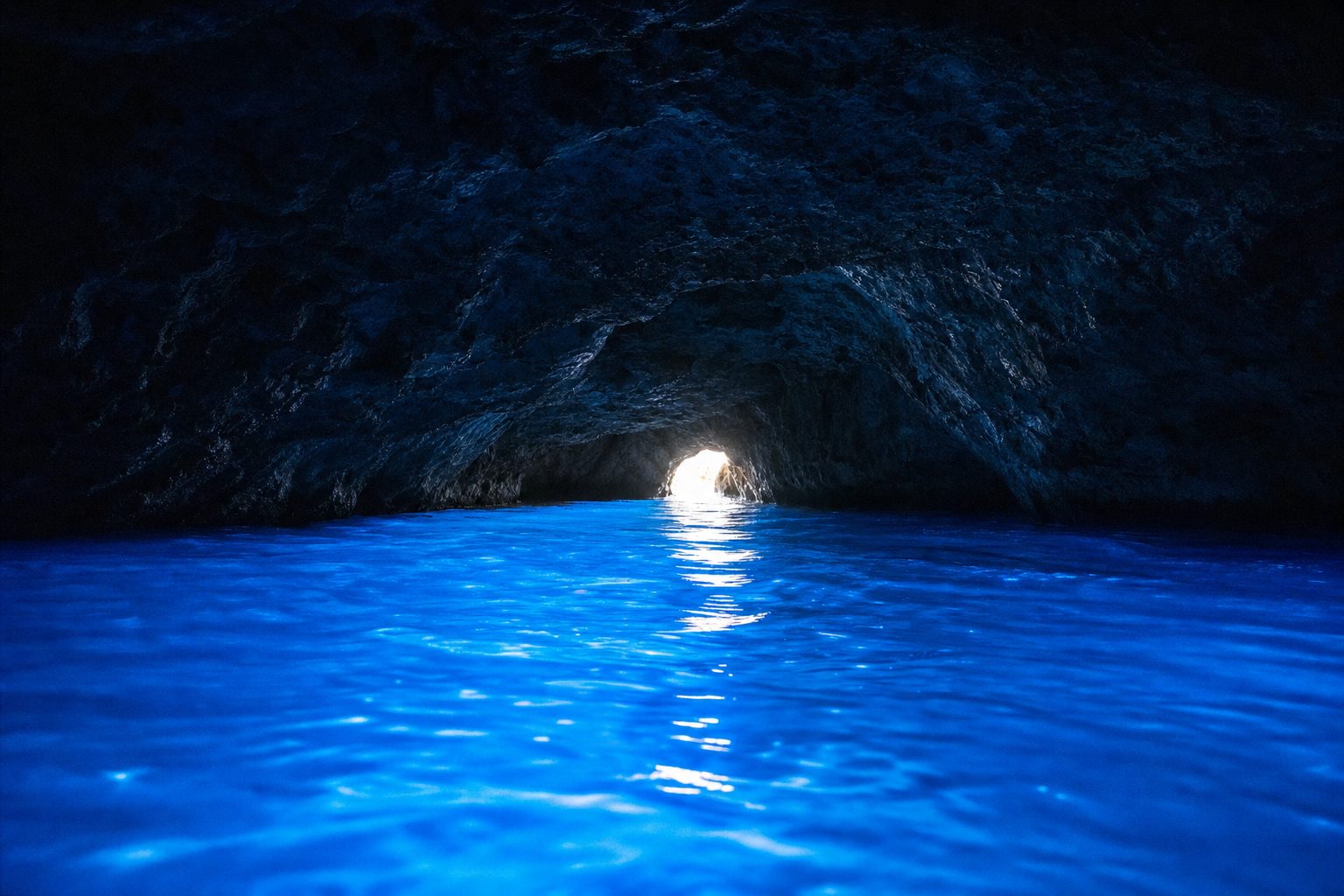 Photorealistic view from dark water inside the Blue Grotto sea cave, electric-blue glowing water in the foreground, the tiny bright entrance arch shining in the midground, and the dark rock vault arching overhead in the background, no people.