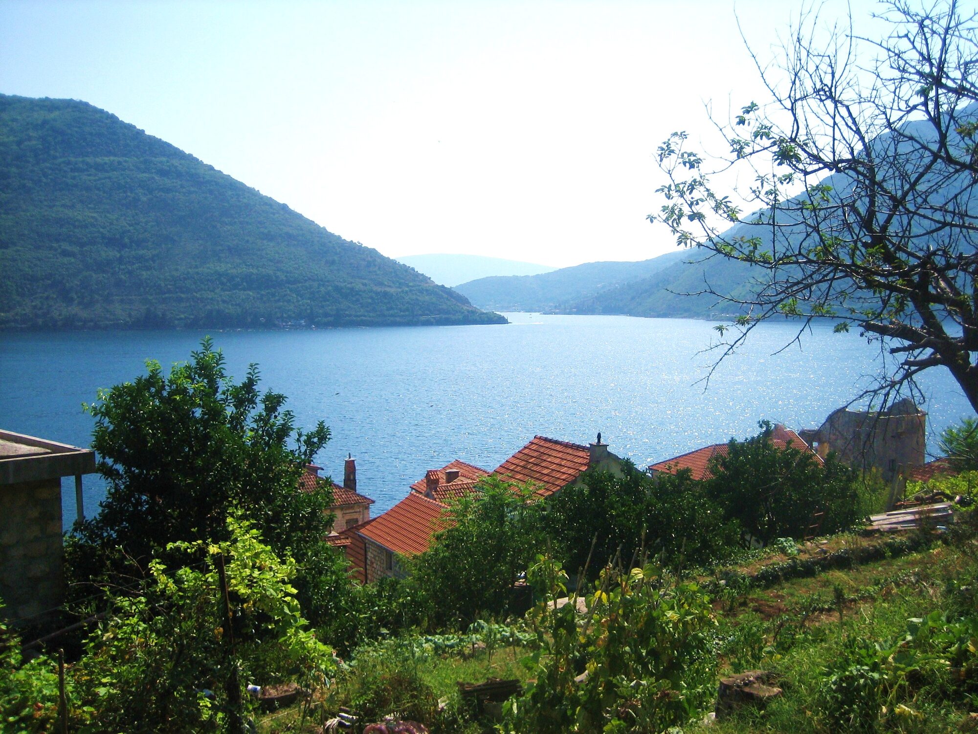 The dark blue Bay of Kotor seen from a hillside staircase in Perast, with steep grey mountains framing the water and red-roofed stone houses along the shore