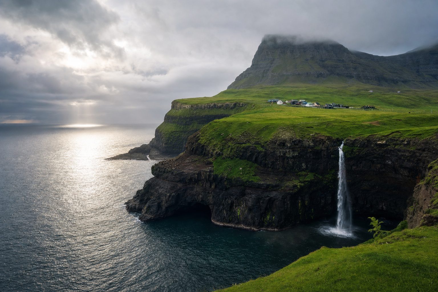 A grass-topped sea cliff village with a single narrow waterfall dropping directly into a churning North Atlantic, small huddled turf-roofed houses in the midground, layered dark headlands fading into mist under a pale overcast sky.