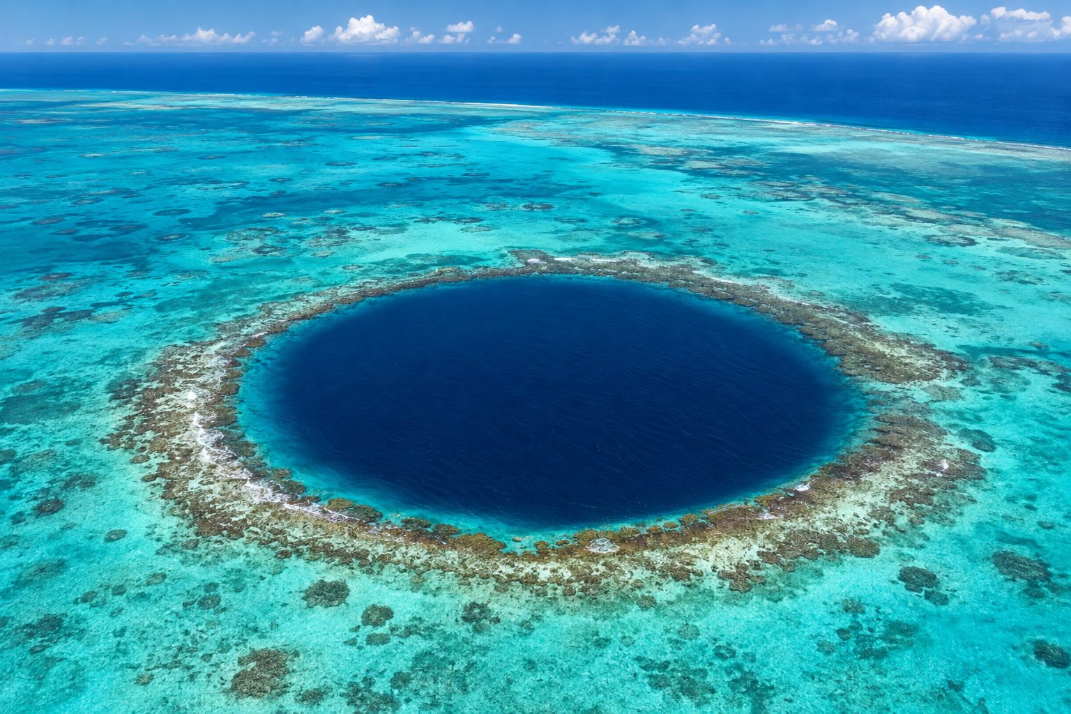 Aerial view of the Great Blue Hole, Belize: a near-perfect circle of deep navy water set in a pale turquoise reef flat, its rim edged by a thin ring of darker coral and white foam, with the shallow atoll lagoon fading to open Caribbean sea and scattered clouds on the horizon under bright midday light.