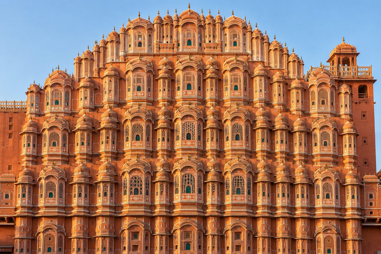 The five-storey pink sandstone facade of the Hawa Mahal in Jaipur fills the frame at golden hour, its hundreds of small latticed windows and domed canopies lit by low warm sunlight, with an empty street in soft shadow in the foreground and a clear pale blue sky above.