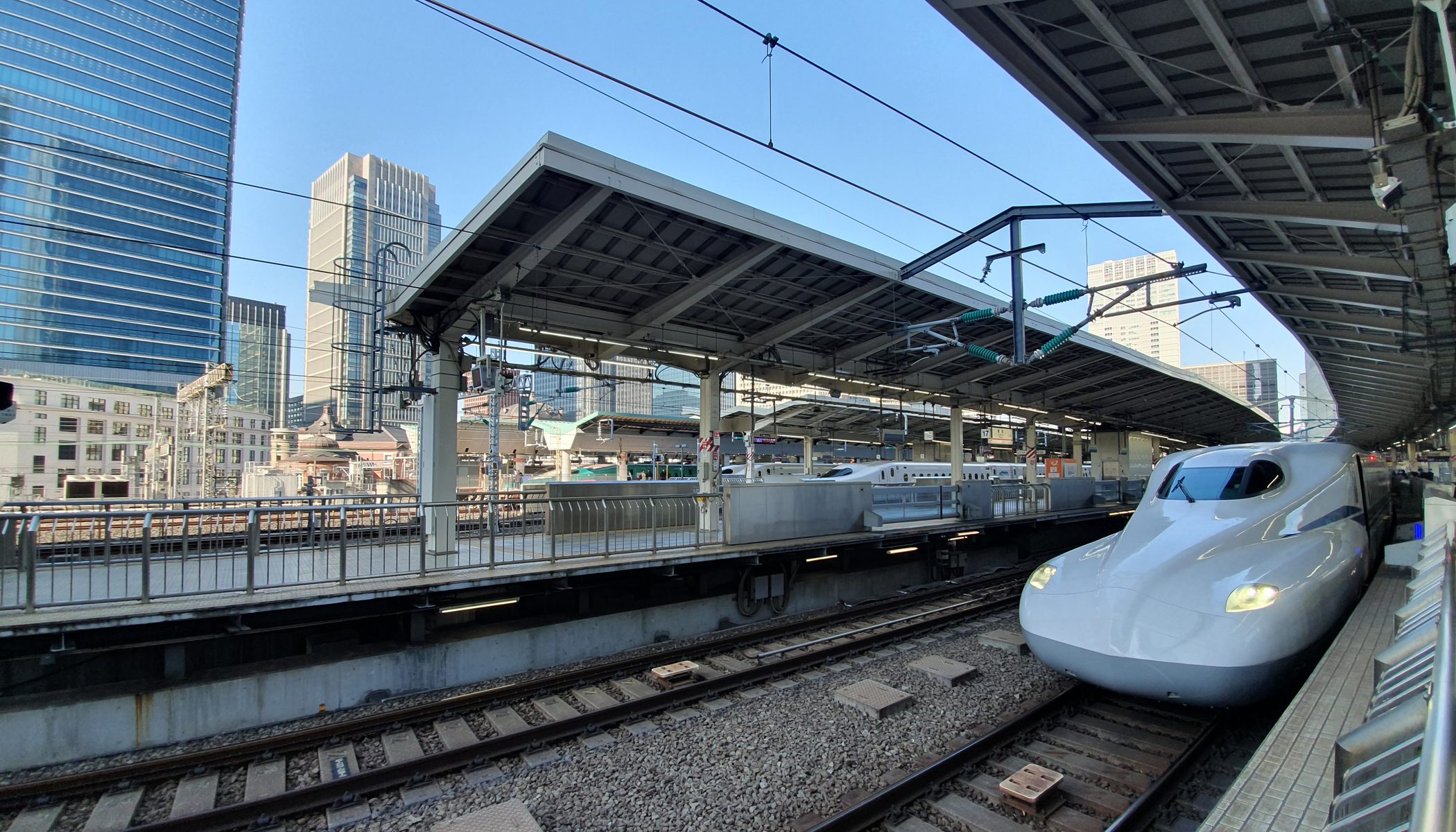 A white Shinkansen bullet train waiting at a covered platform at Tokyo Station, with the city's office towers rising behind the tracks
