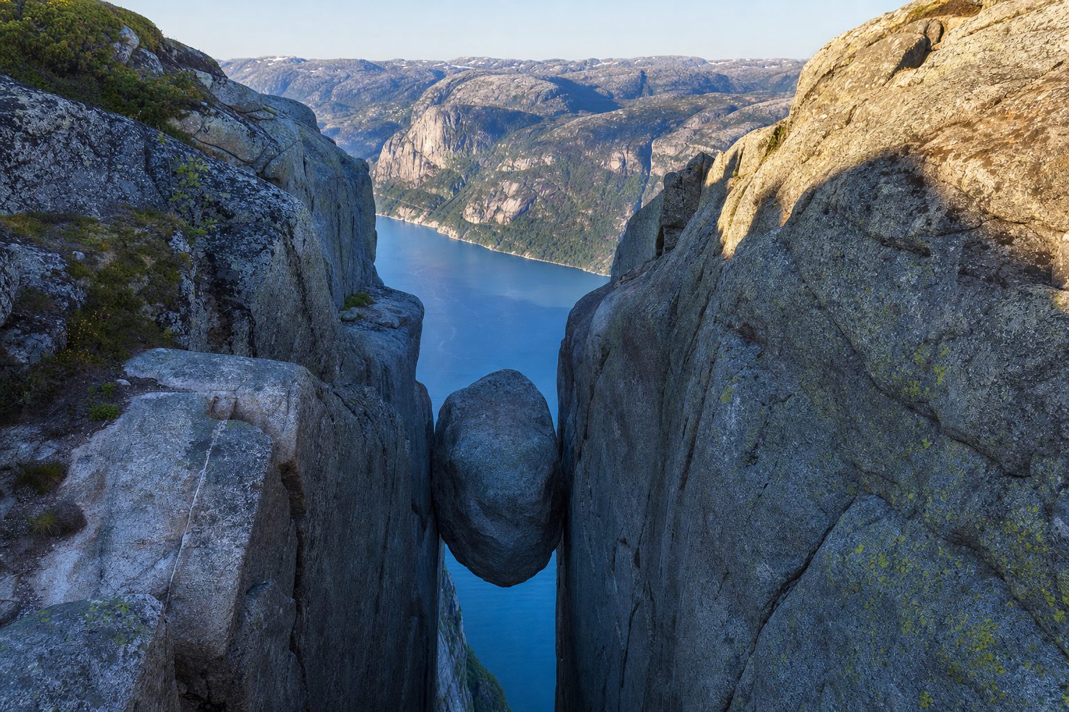 A rounded granite boulder wedged tightly in a narrow crevice between two sheer cliff walls in the foreground, lit by cool morning light; in the midground the cliffs plunge towards the deep blue ribbon of the Lysefjord far below; in the background steep grey fjord walls and layered ridgelines recede under a pale clear sky, with no people visible.