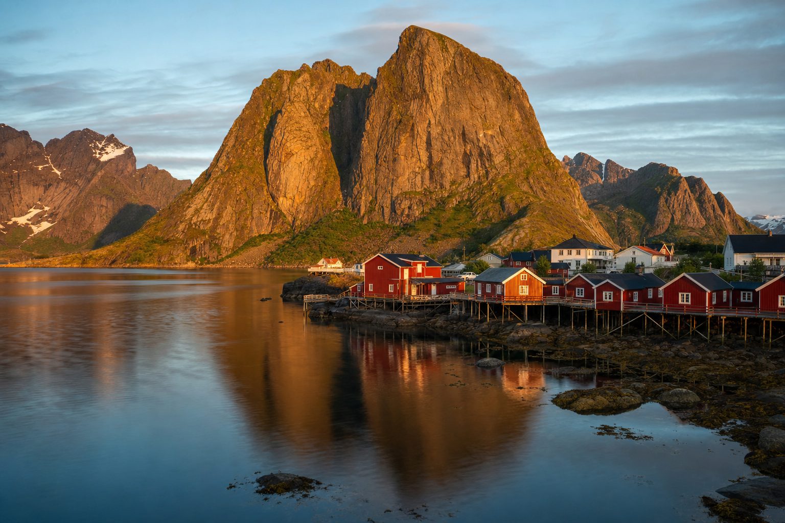 A cluster of red fishermen's rorbu cabins on wooden piles over a still fjord, jagged granite peaks rising sharply behind them under a low golden sun, a narrow gravel road and moored fishing boats in the foreground.