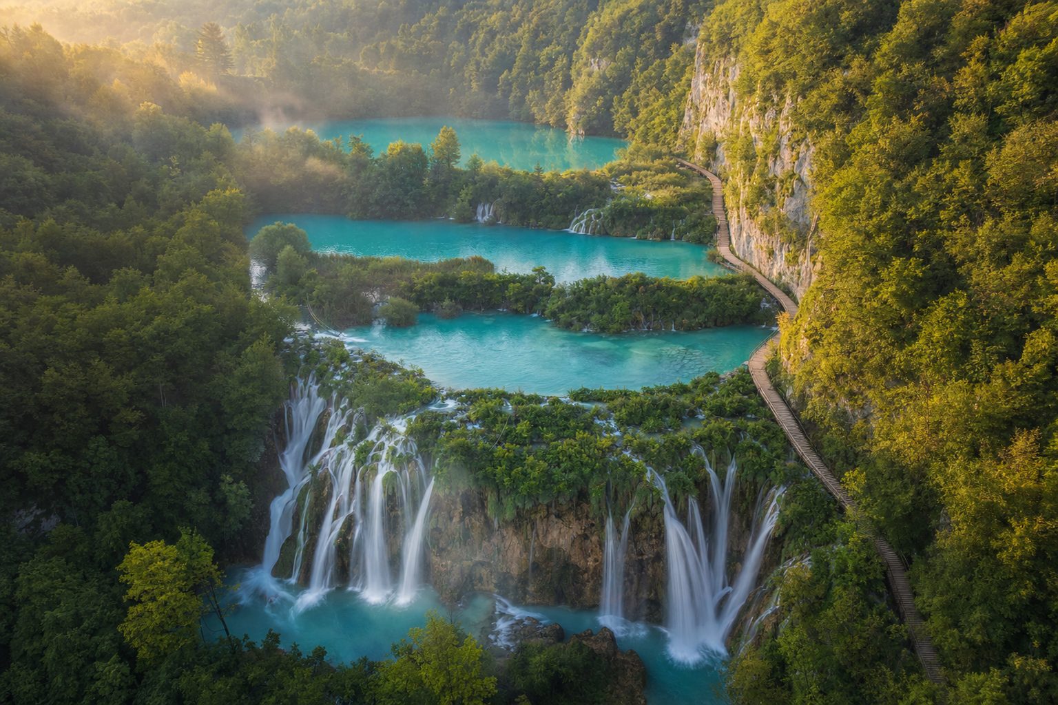 A wooden boardwalk crossing a turquoise travertine lake in the foreground, a tiered waterfall cascading between forested limestone terraces in the midground, and layered green hills fading into soft morning haze in the background, no people visible.