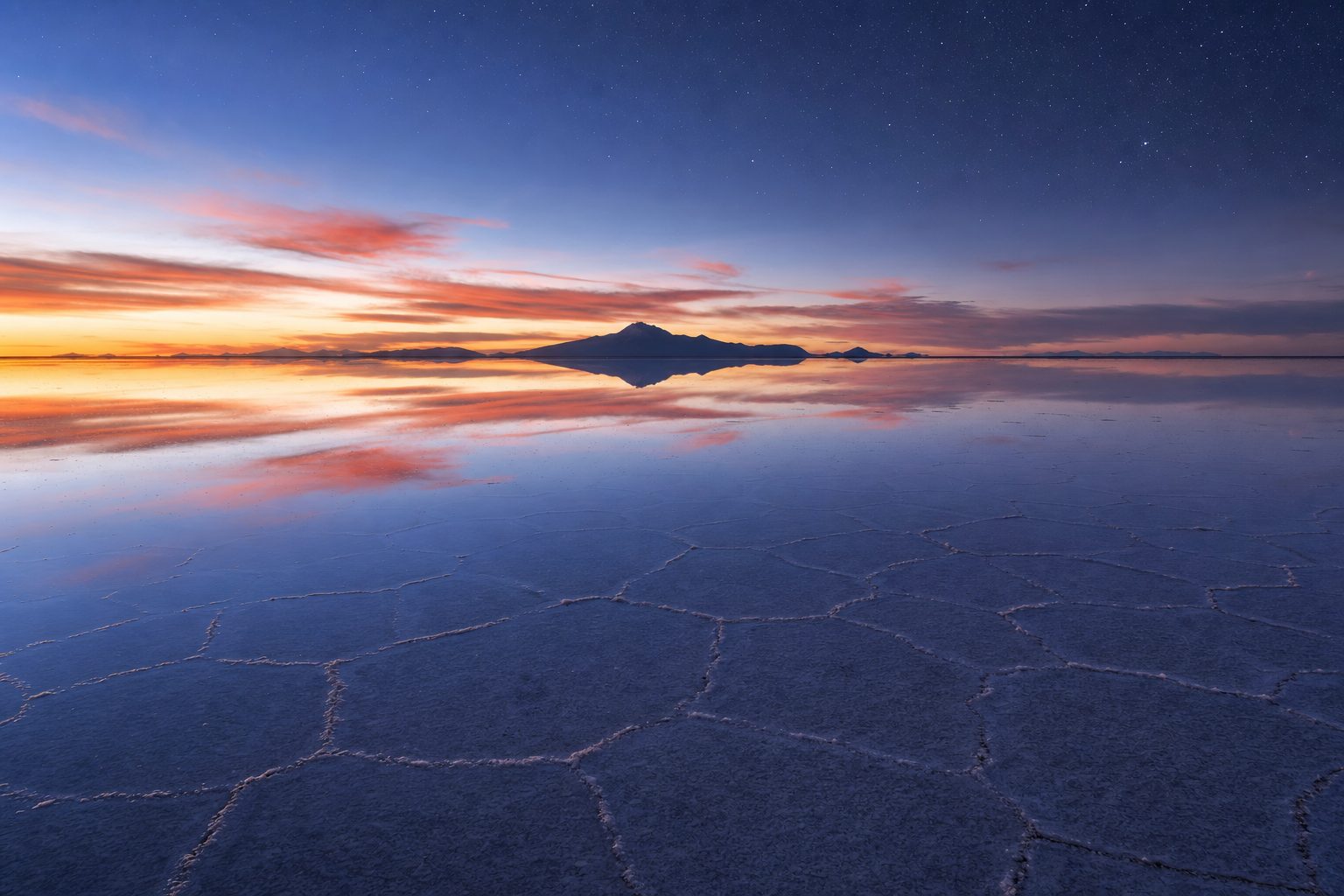 Wide view across the flooded Salar de Uyuni at dusk, with faint hexagonal salt ridges visible beneath a glassy centimetre of rainwater in the foreground, the still surface mirroring peach and violet clouds in the midground, and the dark silhouette of Tunupa volcano doubled in the reflection under a deepening indigo sky with the first stars in the background.