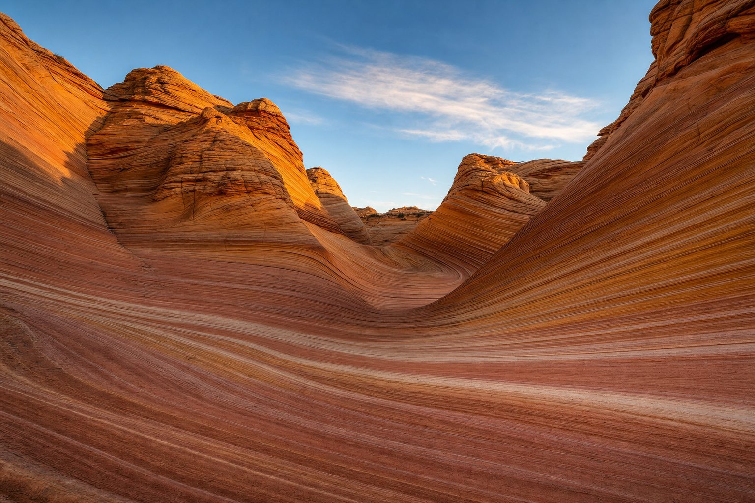 Wide view into the sandstone trough of The Wave in Coyote Buttes North at golden hour, fine rust, salmon and cream cross-bedded stripes curving across the smooth rock floor in the foreground, twisting striped fins rising either side in warm low light with violet shadow in the midground, and a pale blue sky with thin high cloud above the rim of the buttes in the background.