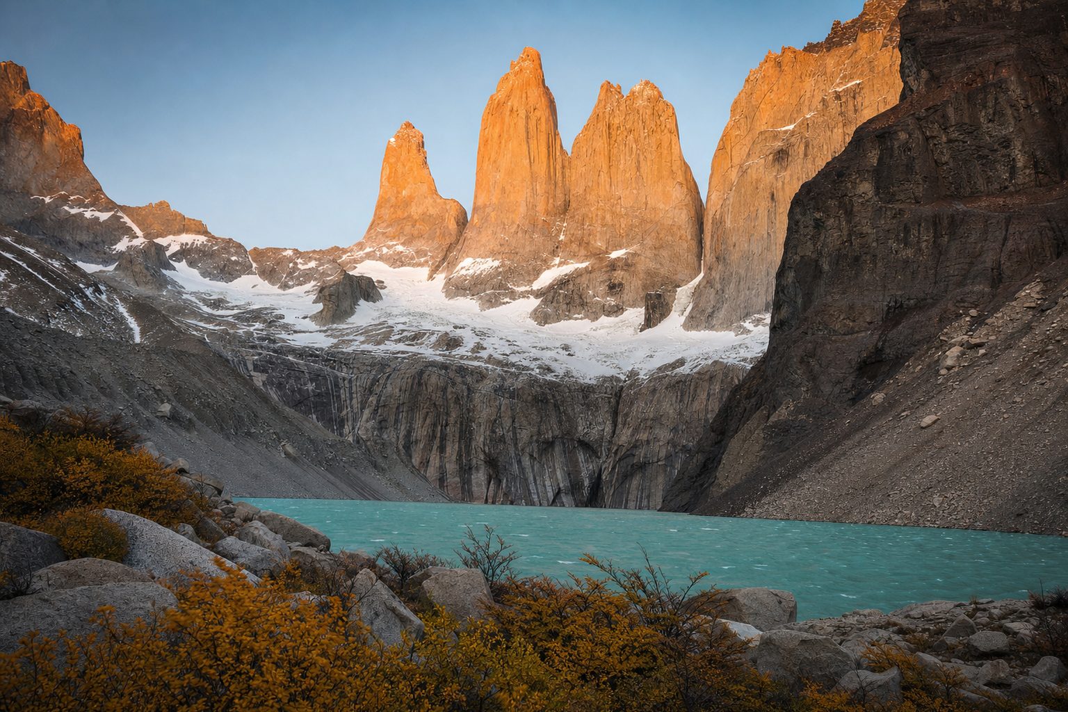 Three sheer granite towers glowing orange at sunrise above a milky turquoise glacial lake, wind-ruffled water and low golden scrub in the foreground, dark scree slopes in the midground, and a clear pale-blue Patagonian sky behind the peaks.