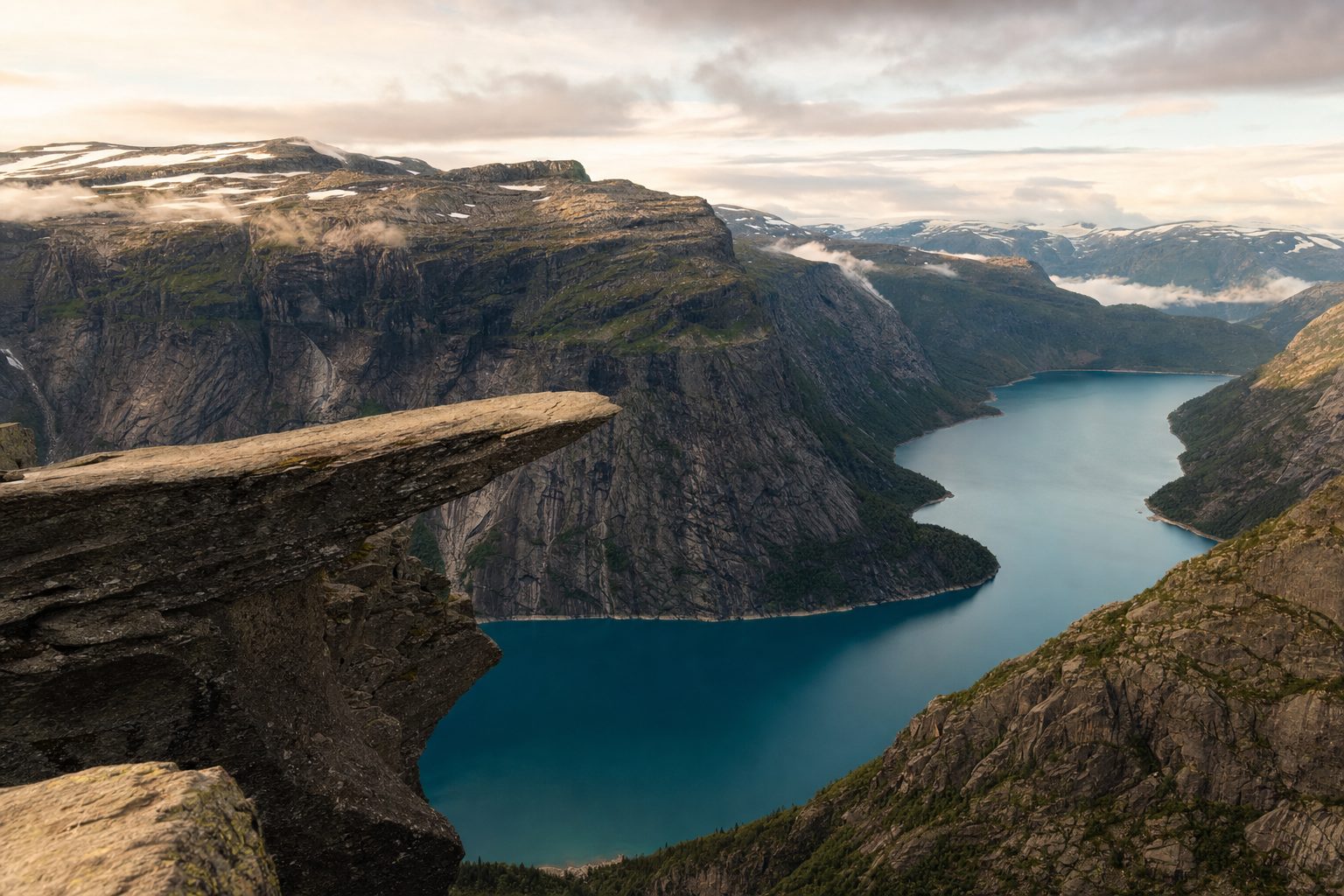The flat granite ledge of Trolltunga projecting from a cliff face in the foreground, the deep blue-green water of Lake Ringedalsvatnet far below in the midground, and steep glacier-carved mountain walls under soft morning light in the background, with no people on the rock.